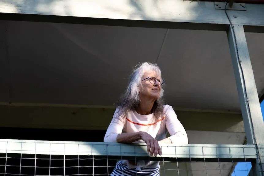A woman with long grey hair and glasses leaning on a white balcony railing gazed into the distance.