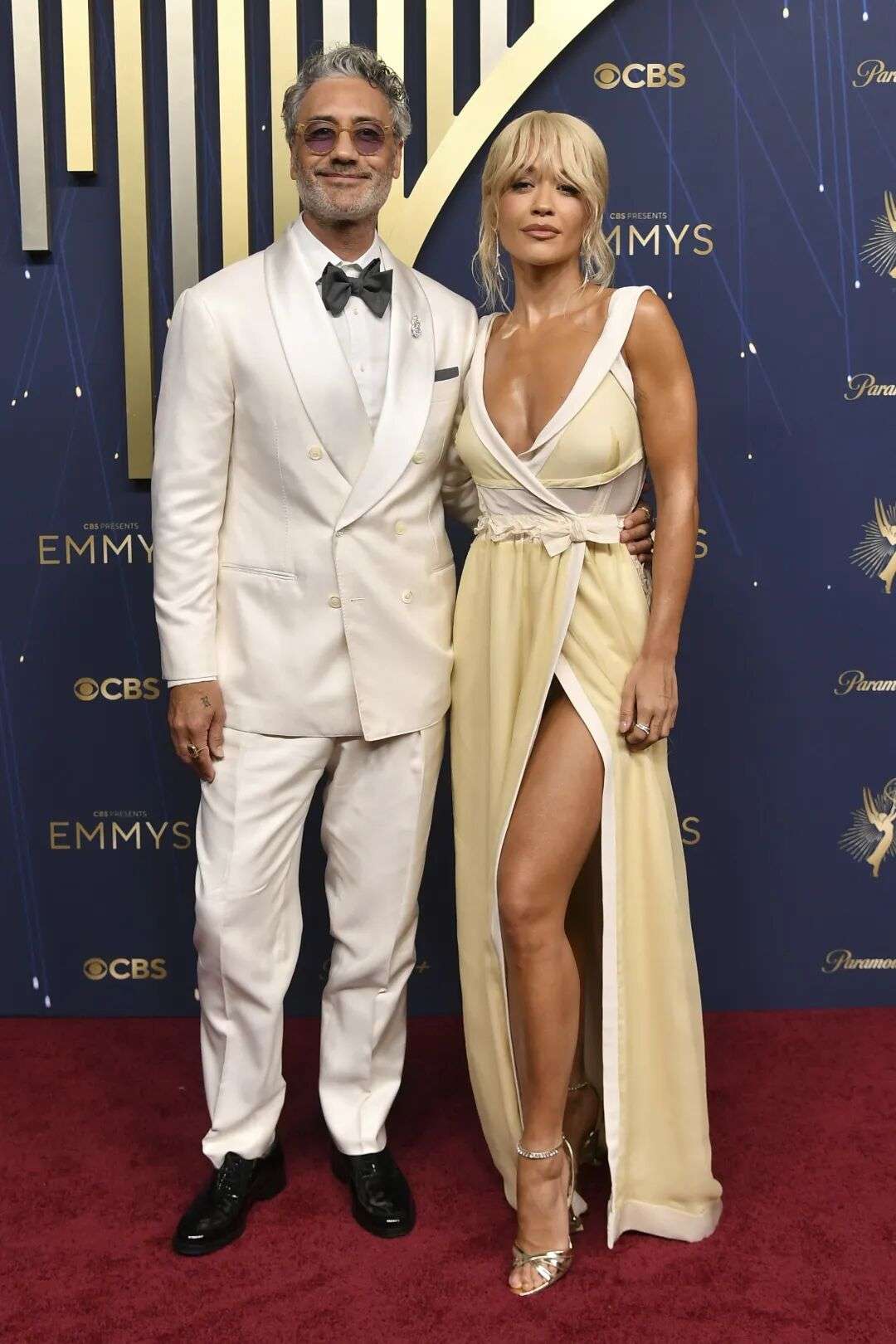 Taika Waititi wearing a white suit and Rita Ora wearing a pale yellow v neck dress with a slit