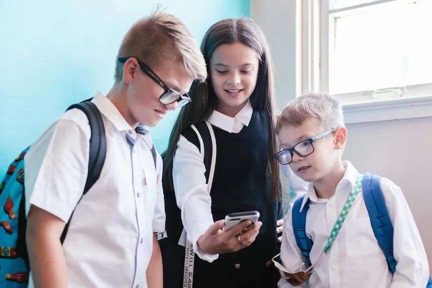 A girl in school uniform shows a phone to two boys in uniform.