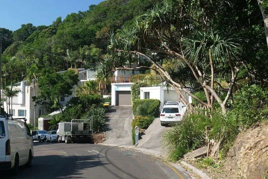 A street lined with cars leads towards several large, modern houses, with dense bush covering the hill behind them.