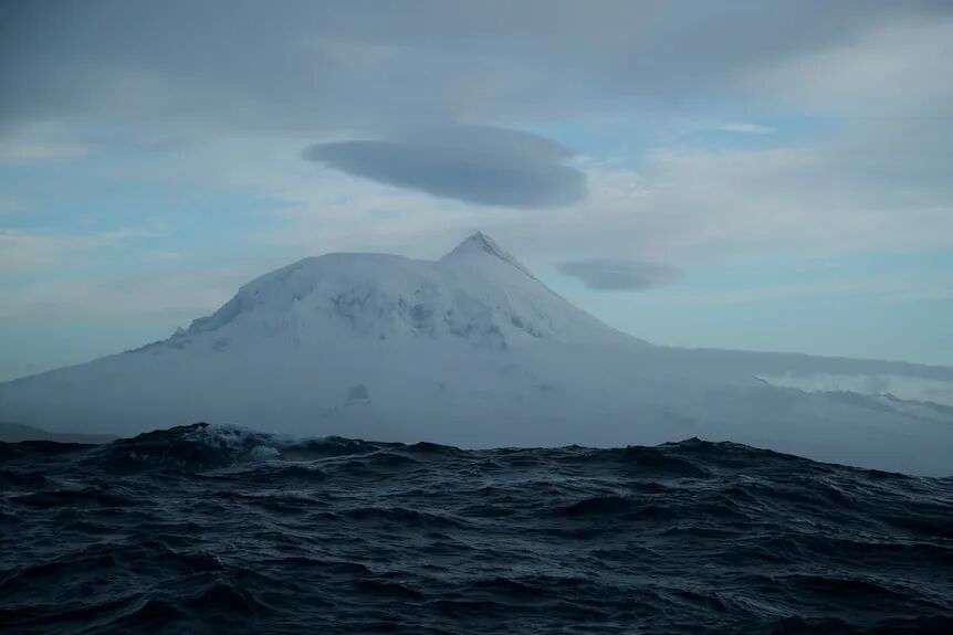 Heard Island and a wave, mountain in the distance, cloud hovering above.