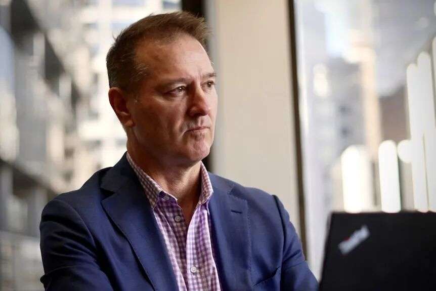 A middle aged man wearing a navy suit jacket and a purple and white checkered shirt.