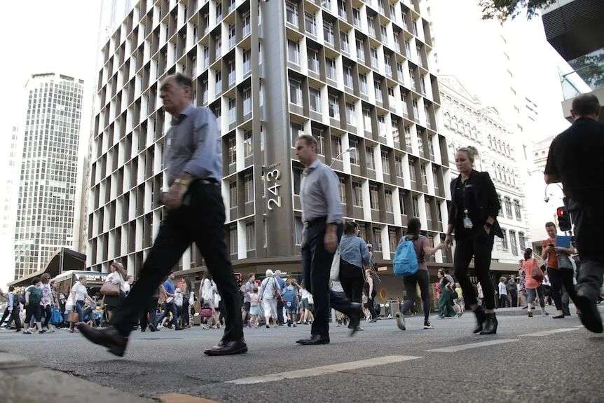 A number of people cross an intersection in Brisbane's CBD. Behind them are office buildings.