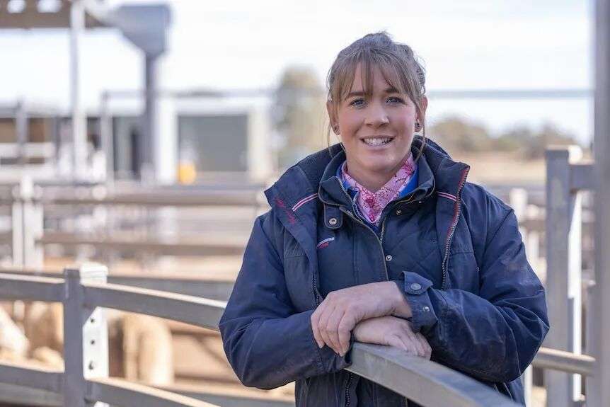 A woman in a shirt and coat smiles while leaning against the fence at a livestock yard