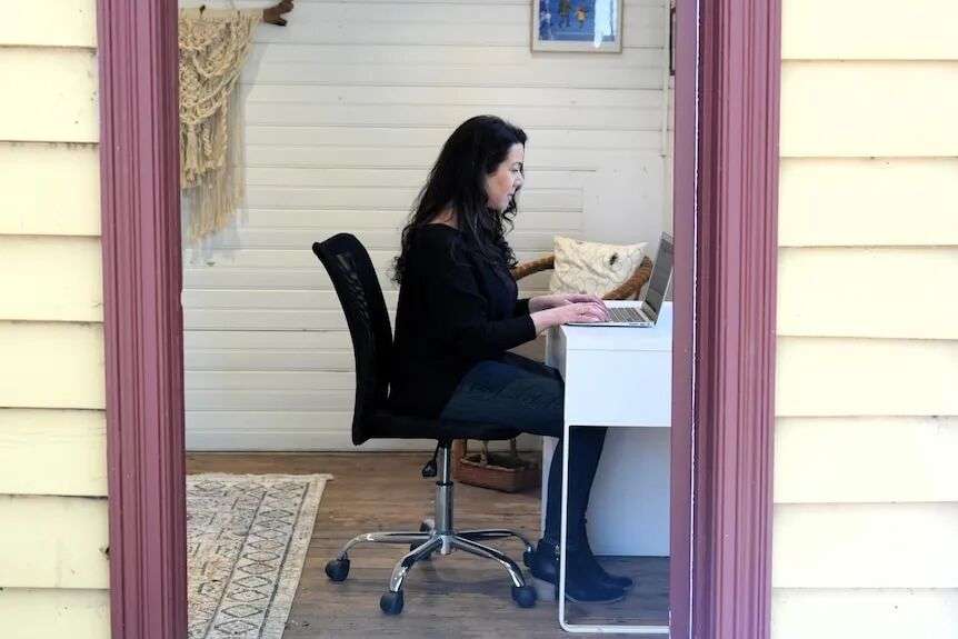 A woman with long black hair and wearing a black top sits at a laptop at a desk.