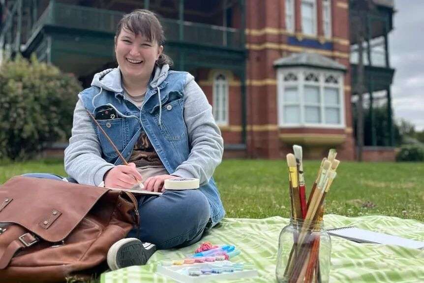 A young woman in a grey hooded jumper and denim jacket sits on a rug on a lawn with a paint brush in hand.