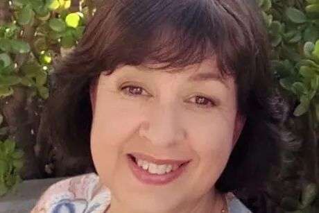 A woman with brown eyes and brown hair wearing a necklace and blue jumper with pink and beige floral designs.