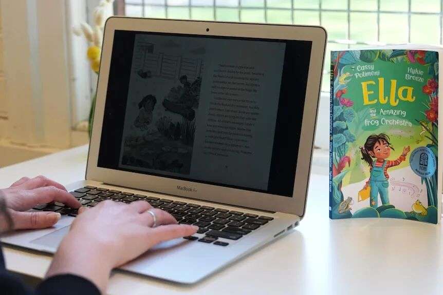 An image focusing on Cassy's hands typing on a laptop next to a colourful green children's book at a desk.