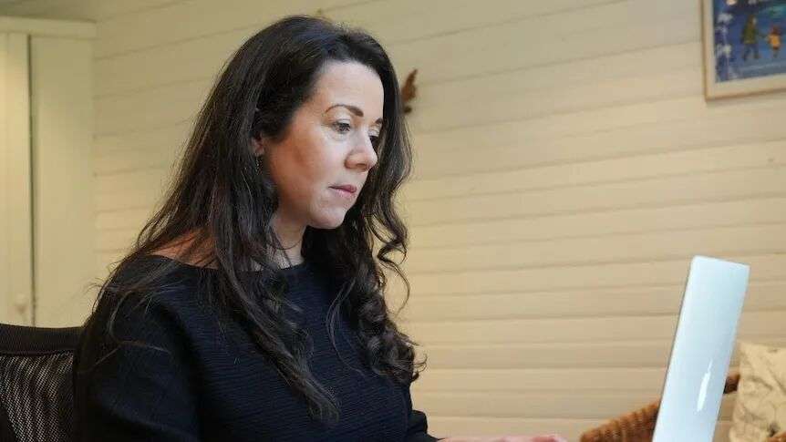 A woman with long black hair and wearing a black top sits at a laptop at a desk.