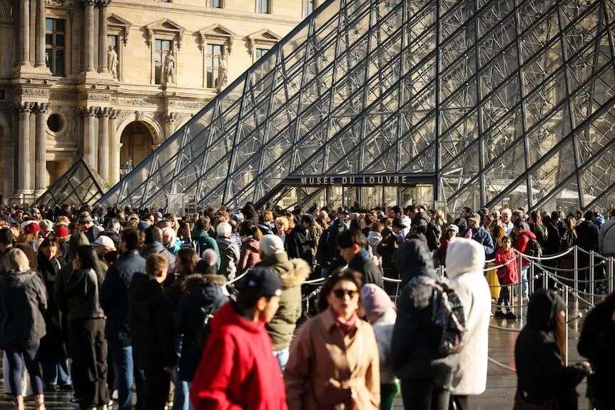 Visitors queue outside the Louvre museum on a sunny day.