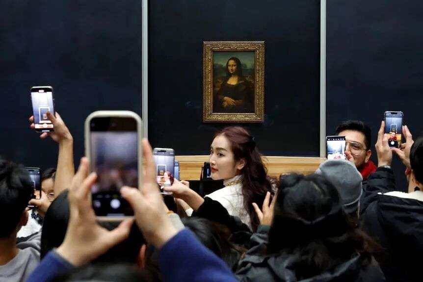 A crowd of visitors take photos and stare at the Mona Lisa painting hanging on a wall at the Louvre.