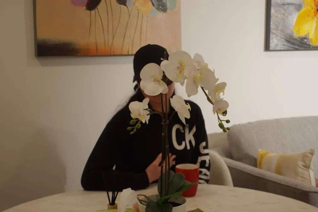 A person wearing a black cap and sweatshirt sits at a table, her face mostly obscured by a white orchid.