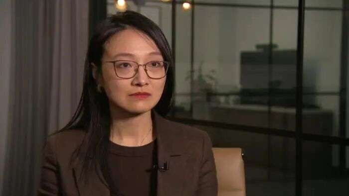 A woman wearing glasses and a brown blazer over a brown top, looking directly at the camera in a warmly lit, modern indoor setting.