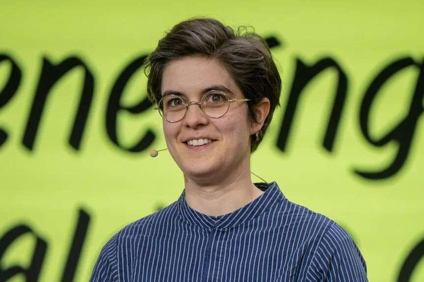 Marlene Engelhorn smiles at the camera. She is wearing round reading glasses. And a blue top.