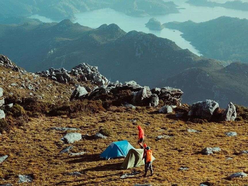 An aerial view of two people standing next to hiking tents near a rocky outcrop overlooking a lake down below.