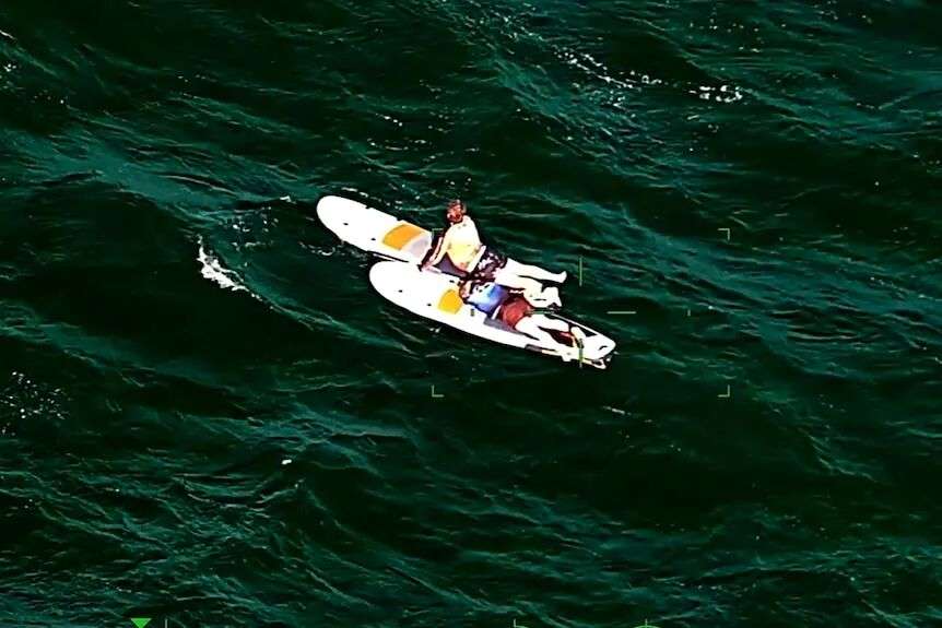 An aerial photo of two people lying on paddleboards on a green choppy sea.