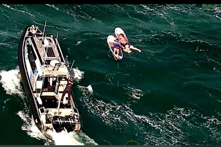 A police boat near two people lying on paddleboards in a green choppy sea.