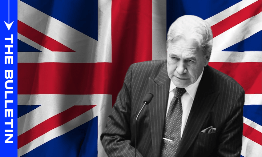 A man in a suit speaks at a podium in front of a large British flag; the left side has a blue vertical banner with the words 