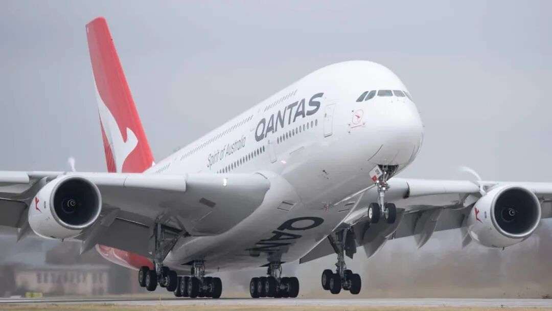 A photo of a Qantas Airbus A380 in the airline's famous white and red livery takes off from an unknown airport. The plane has just rotated for ascent, its nose in the air and its rear wheels still on the ground. Heat from its engines radiates from the back of its wings against a grey sky.