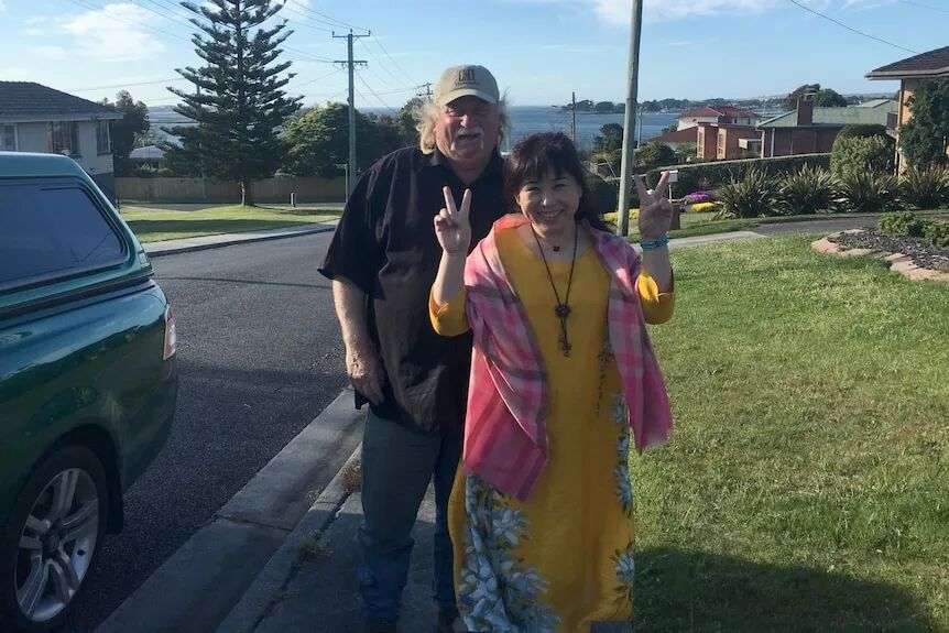 A man with white hair and a baseball cap smiles and stands behind a smiling woman throwing up the peace sign with her hands.