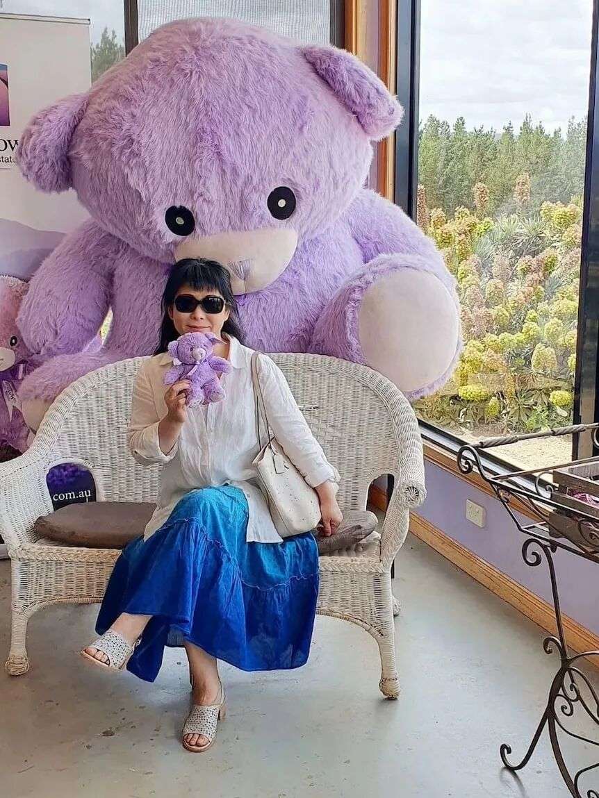 A woman in sunglasses sits on a seat and holds a purple teddy bear.