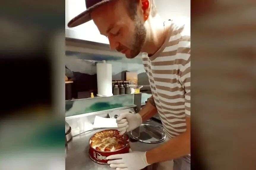A young man with a short beard cuts a cake in a commercial kitchen.