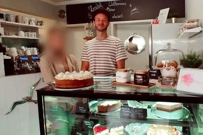 A young man and a woman stand behind a cake counter in a cafe.