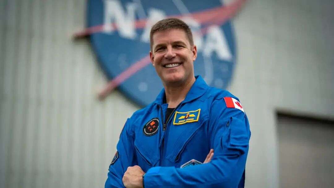 A man in a blue astronaut jumpsuit stands with his arms crossed in front of the blue NASA logo.